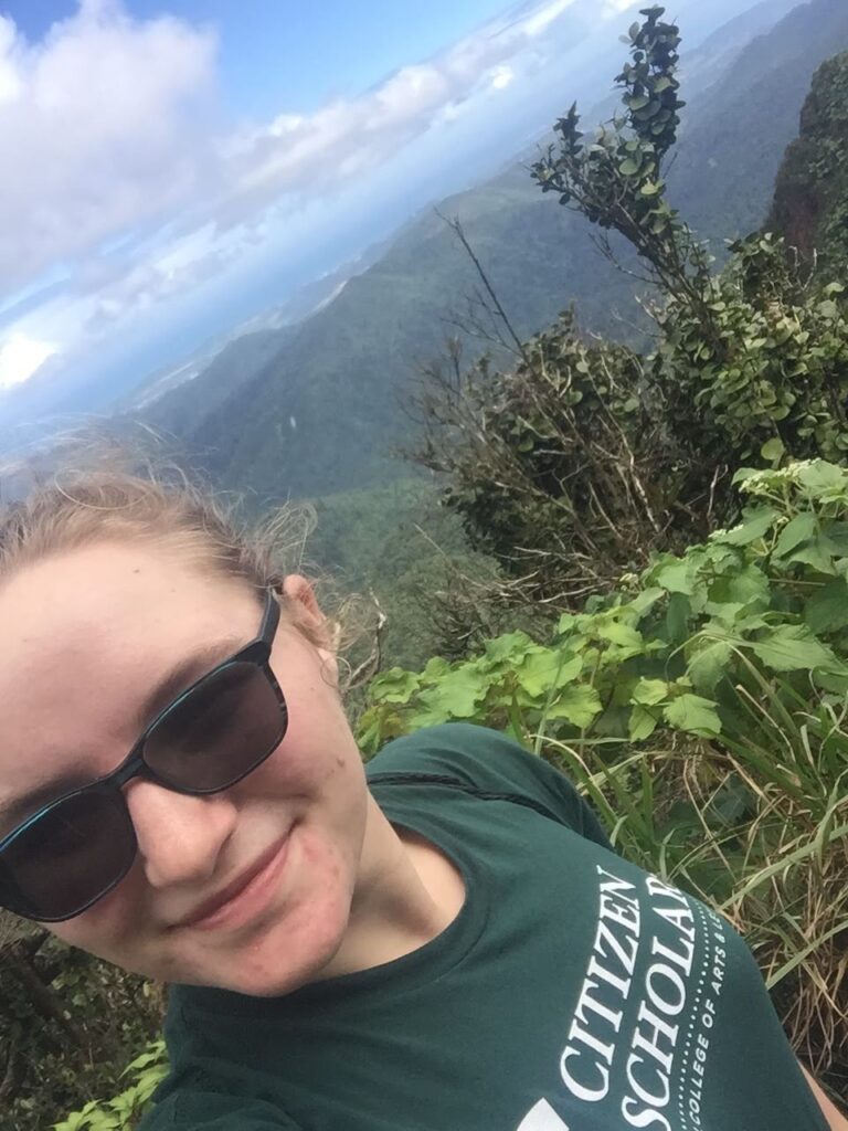 A woman stands in front of a rainforest with sunglasses.