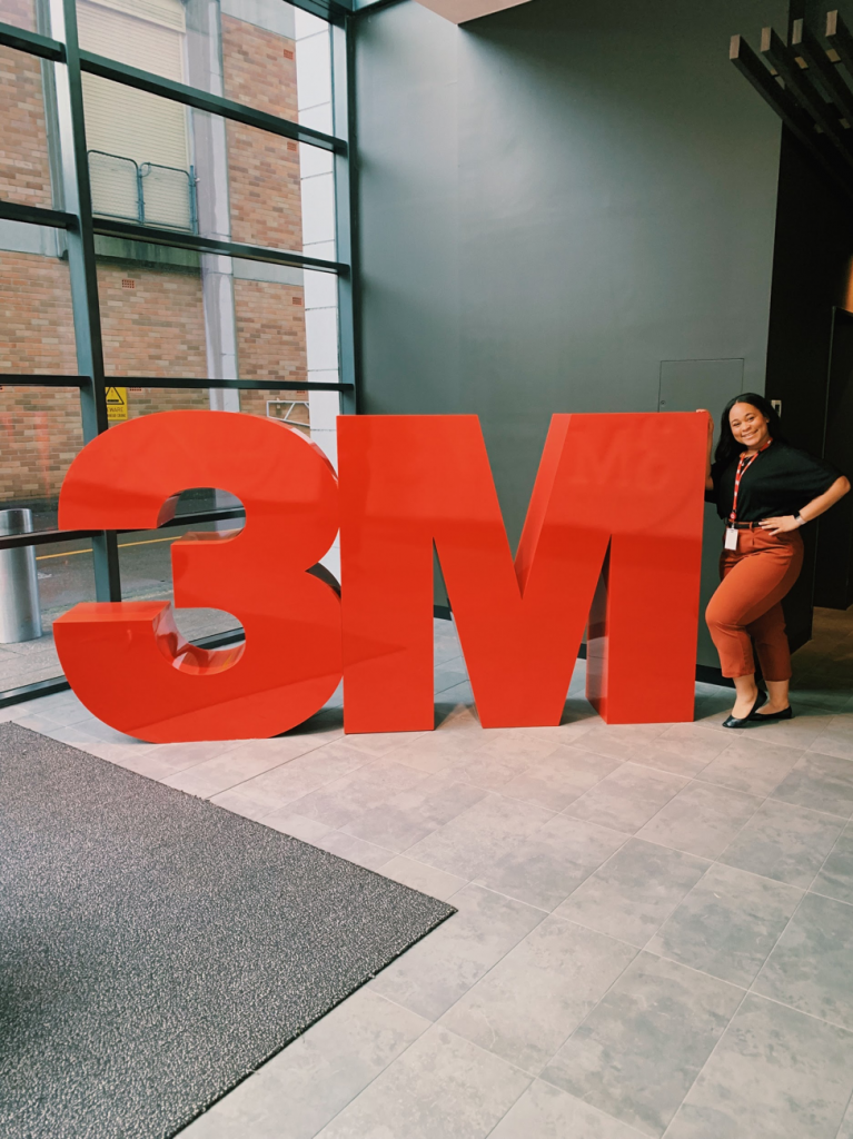woman standing next to giant "3M" logo in office space. 
