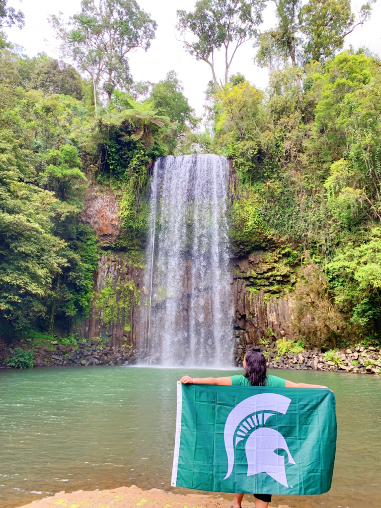 woman standing in front of waterfall with back to camera, holding green flag with spartan logo. 