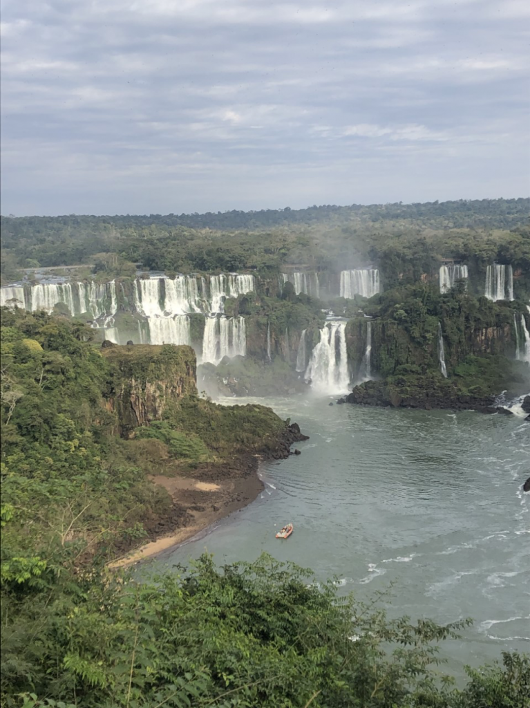 series of waterfalls coming from a thick forest and a large lake with a small boat on the water