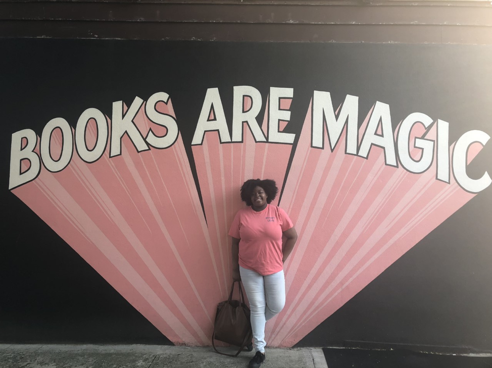 Black woman wearing pink shirt standing in from of street art that reads "books are magic"