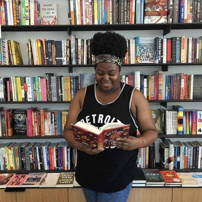 Black woman standing in front of bookshelf smiling and looking down at book in hands