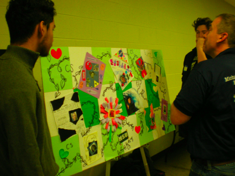 Two students are standing to the left and right of their project, as a person looks at the poster. 