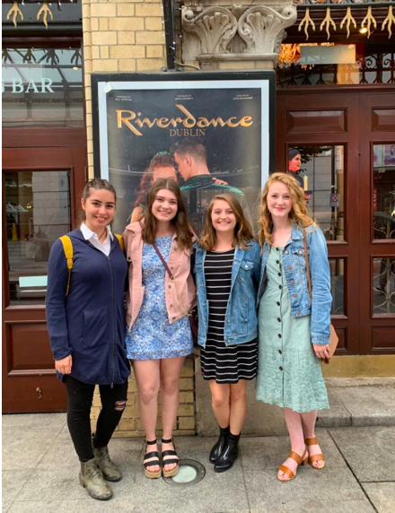 Four women stand in front of a poster that says, "Riverdance." Behind the poster are two brown doors.