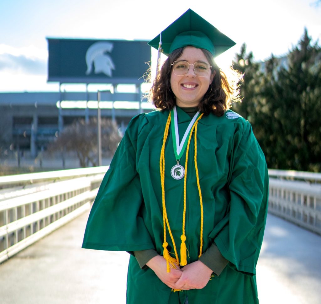 Photo of person standing outdoors on a bridge in front of a football stadium. The person is in a green graduation gown.