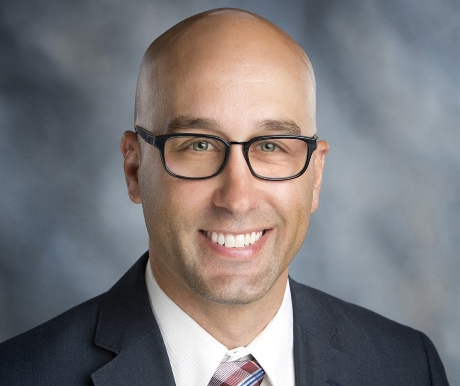 Photo of a man who is bald, smiling and wearing glasses and a suit and tie. 